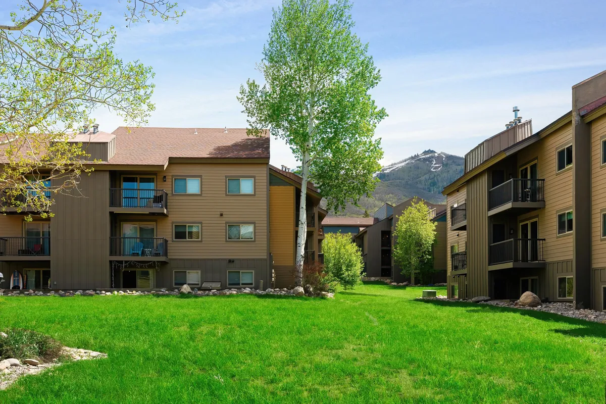 A broad exterior view of the property with green grass, tall trees, and mountains as a backdrop. The balconies of the apartments are visible, indicating spaces for both private and communal outdoor enjoyment. The overall mood is serene and communal, inviting guests to relax or gather outdoors.