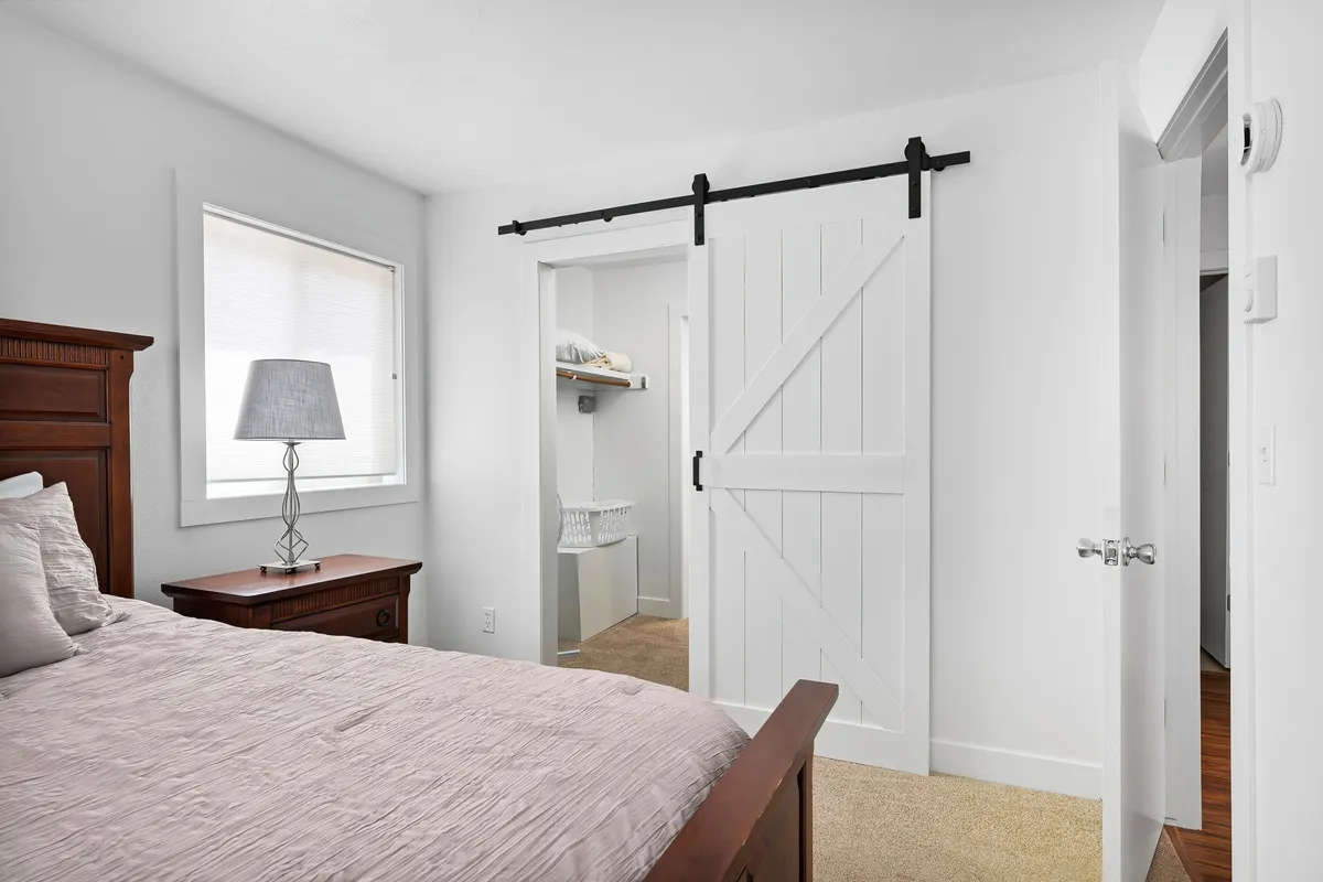 This image features a serene bedroom with a plush bed adorned with textured bedding, a bedside table with a lamp, and a barn-style sliding door. Soft natural light filters into the room, enhancing the peaceful atmosphere.