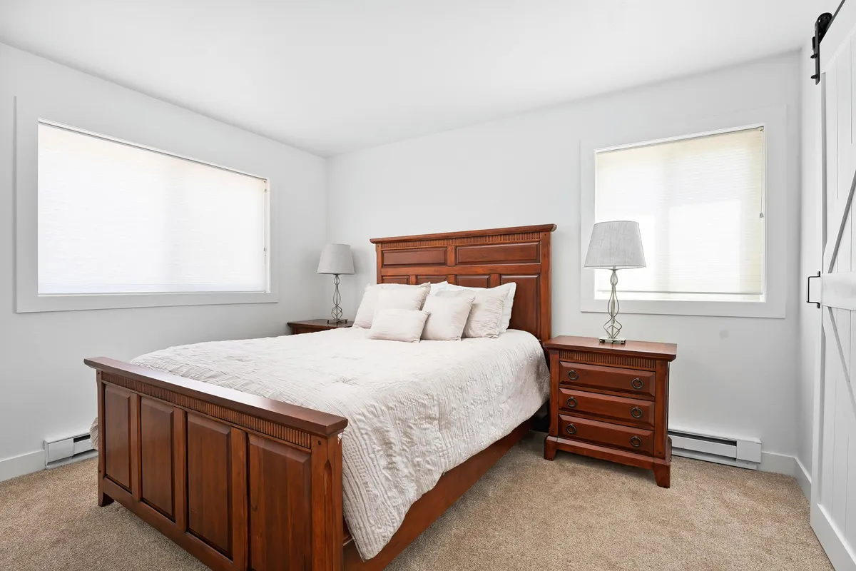 A cozy bedroom with a wooden bed frame, neatly arranged bedding, nightstands, and lamps. The scene is filled with soft, warm light, offering a sense of sanctuary and relaxation.