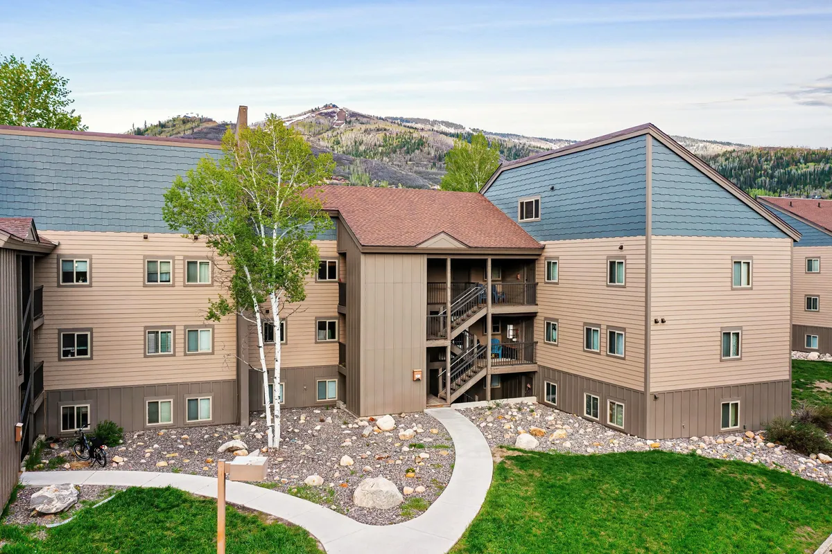 A multistory building sits amid a landscaped pathway and rock garden, with surrounding mountains in the distance. The inviting pathway suggests convenience and a welcoming atmosphere for returning after snowy adventures.
